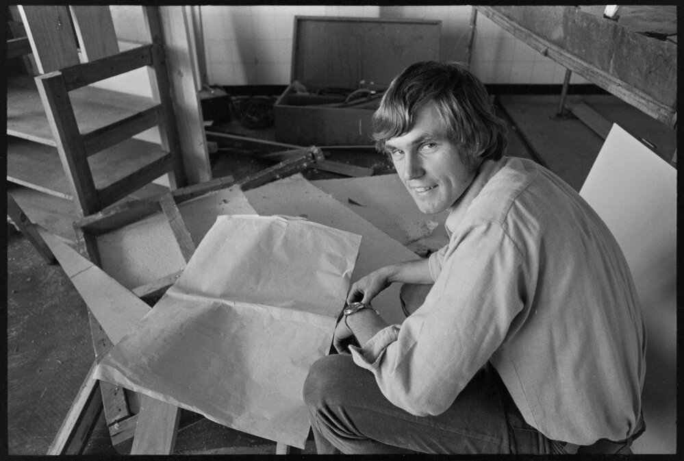 Black and white photograph of Mr Trevor Gregory crouched down in his new butcher shop being renovated, reviewing plans